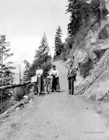 Two women bicyclists standing next to their bikes and a man without a bike posing on Chuckanut Drive, ca 1916.